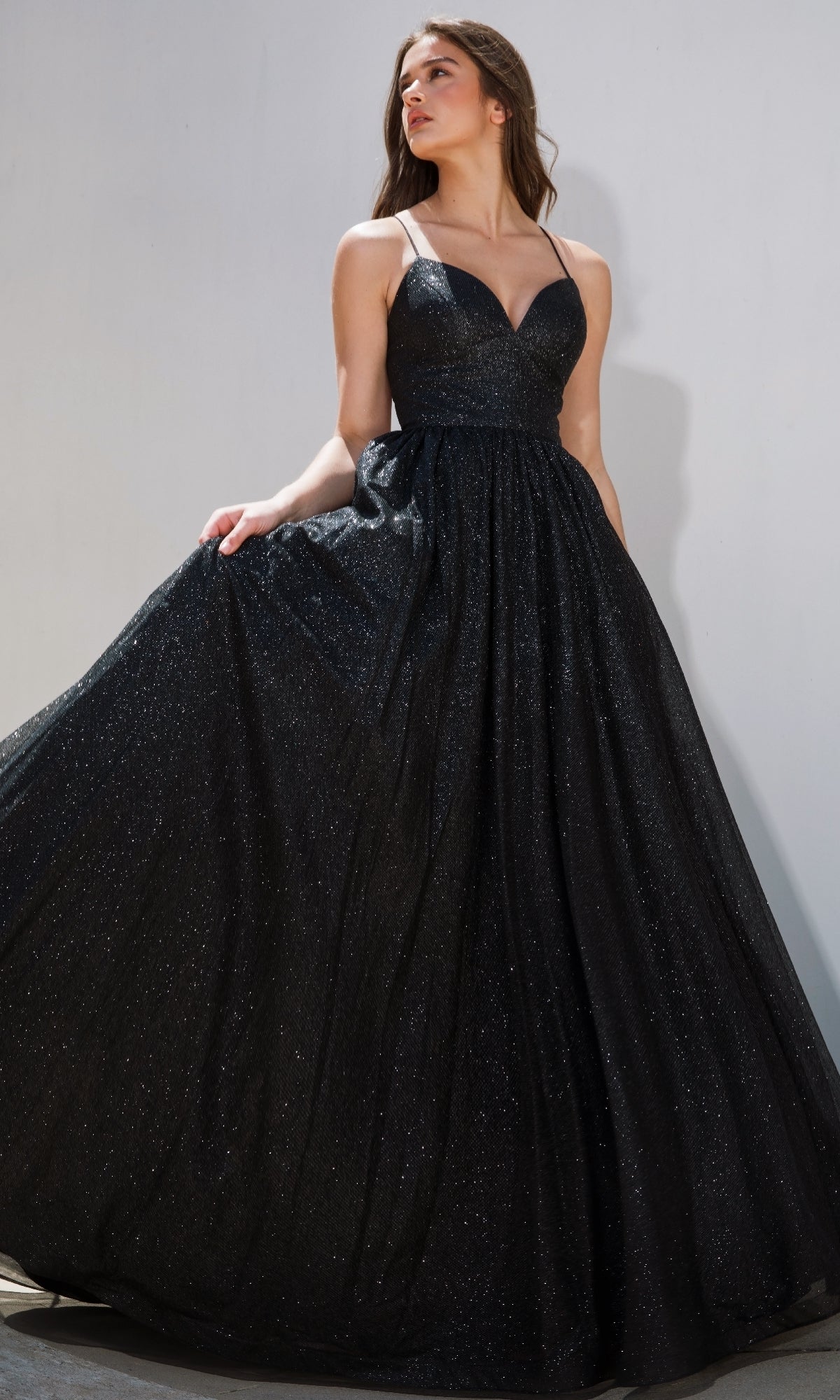 A woman poses against a plain light background wearing the Strappy-Back Glitter Prom Ball Gown: Eureka 9009, a sleeveless, floor-length black gown with a deep neckline and sparkly fabric.